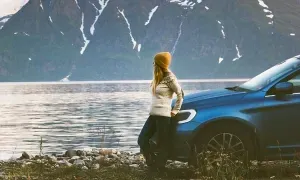 vrouw met muts leunt tegen een blauwe auto aan de rand van een meer met een berg op de andere oever