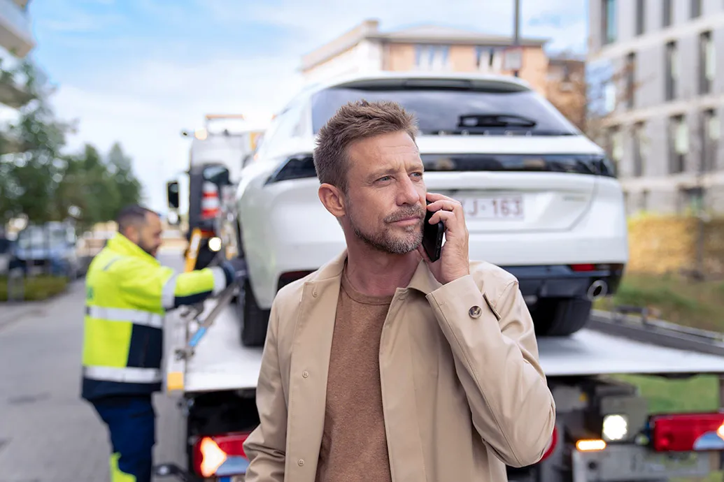 man van middelbare leeftijd telefoneert terwijl hij voor een takelwagen staat waarop zijn auto bevestigd wordt door een VAB-wegenwachter