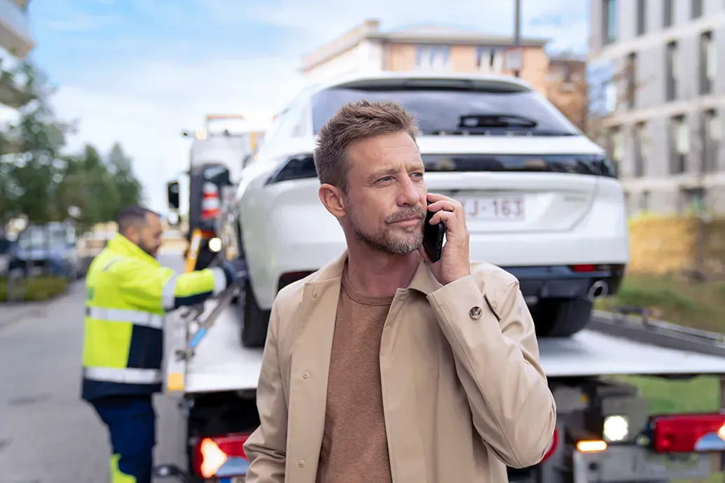 man van middelbare leeftijd telefoneert terwijl hij voor een takelwagen staat waarop zijn auto bevestigd wordt door een VAB-wegenwachter