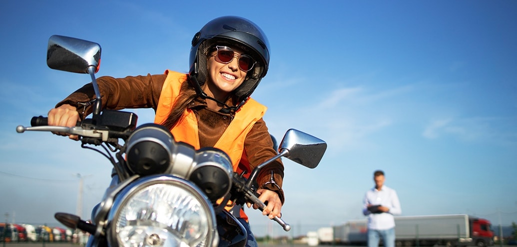 meisje op een moto lacht tijdens een rijles, op de achtergrond zie je een instructeur