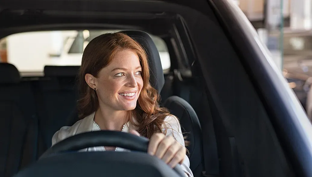 een vrouw met rood haar lacht en zit achter het stuur van een auto met een donker interieur