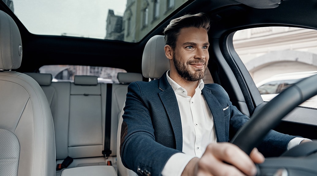 een man met korte baard, kostuum en hemd zit achter het stuur van een auto met een lichtgrijs interieur