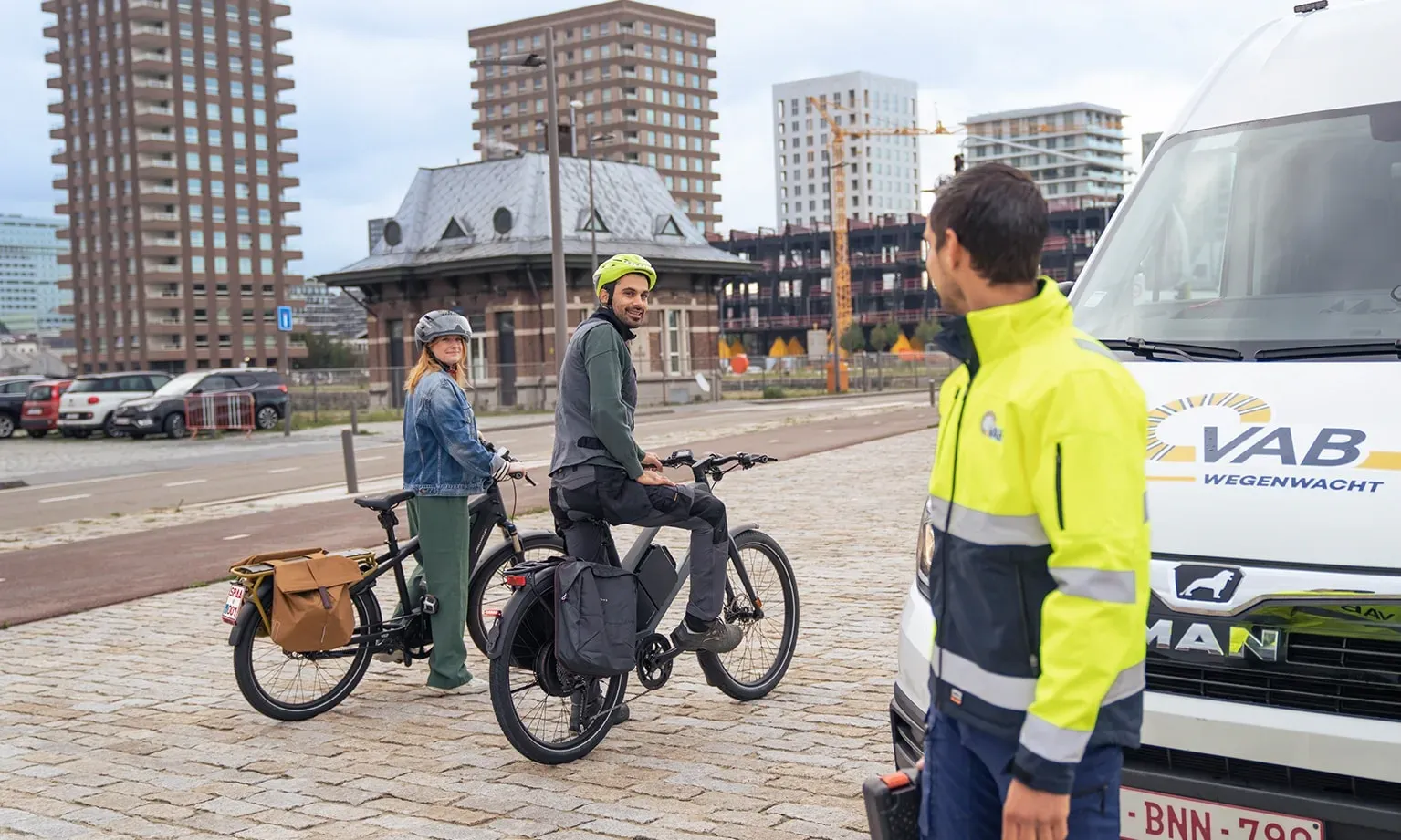 Wegenwachter staat bij een VAB‑voertuig terwijl twee personen met hun fietsen stoppen op een plein