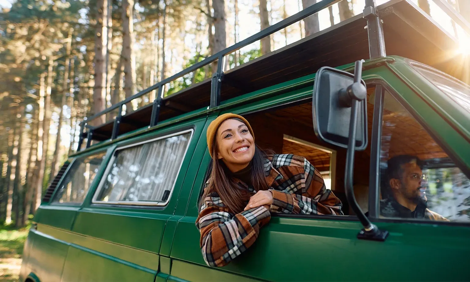 Femme dans un van aménagé se penche par la fenêtre tandis qu’elle est garée avec son partenaire dans un paysage forestier.